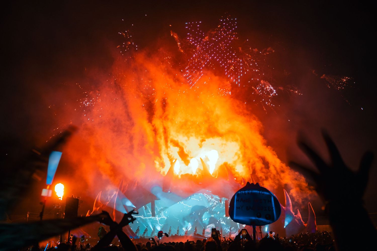 Concert crowd silhouettes against vibrant orange and blue pyrotechnic display during nighttime music festival performance.