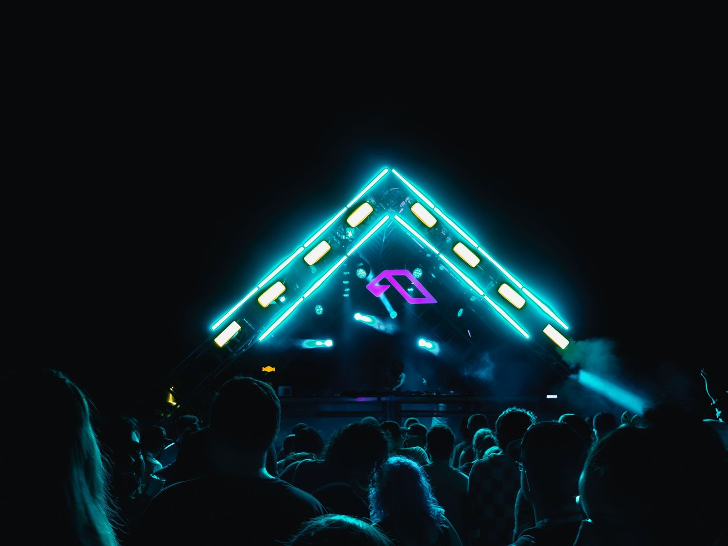 Illuminated triangle stage with neon blue lights hovers over a dark silhouetted crowd at an electronic music concert.