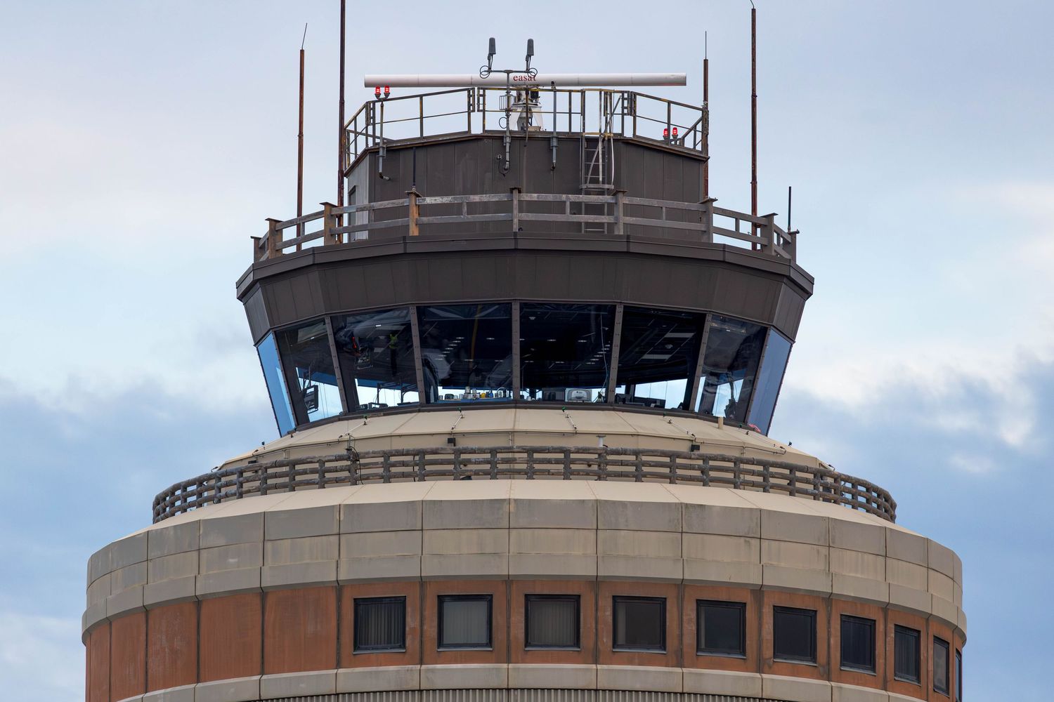 Close-up view of an airport air traffic control tower with large windows and communication antennas against a cloudy sky.