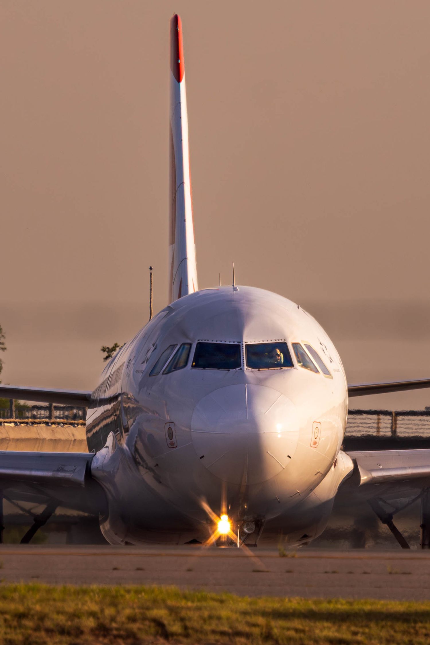Commercial airplane with red tail livery taxiing on runway at golden hour sunset, front view with landing light on.