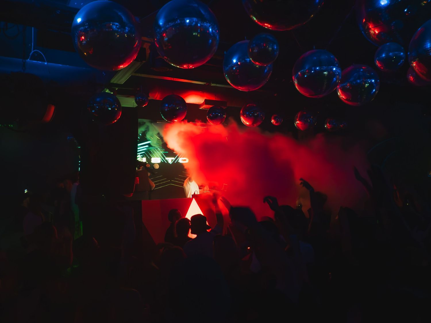 Nightclub interior with glowing disco balls overhead and dramatic red lighting illuminating a smoky atmosphere.