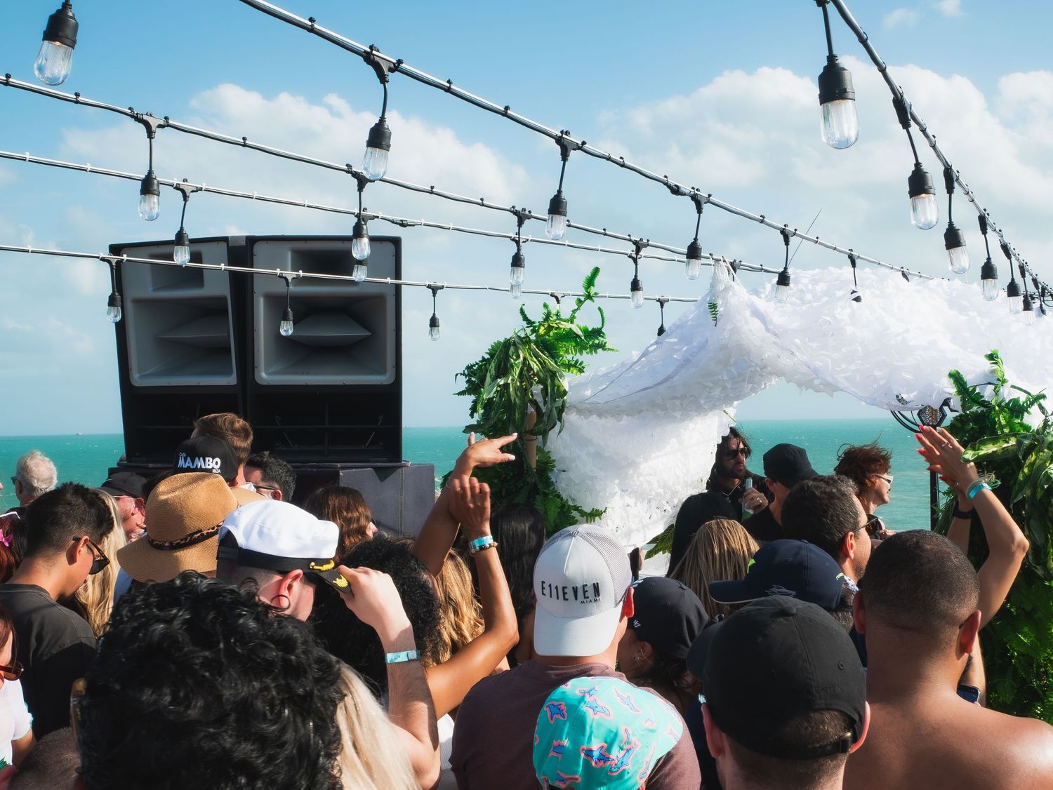 A crowd dances at a beach party under string lights with speaker stacks and an ocean view in the background.