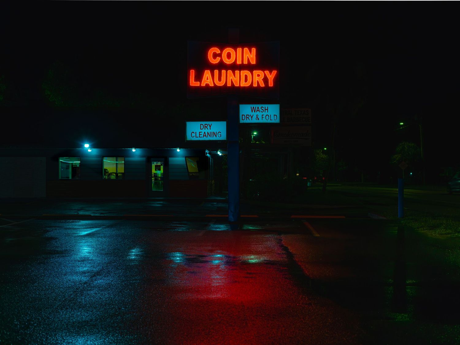 Neon red 'COIN LAUNDRY' sign glows in the dark above a wet parking lot at night, casting colorful reflections.