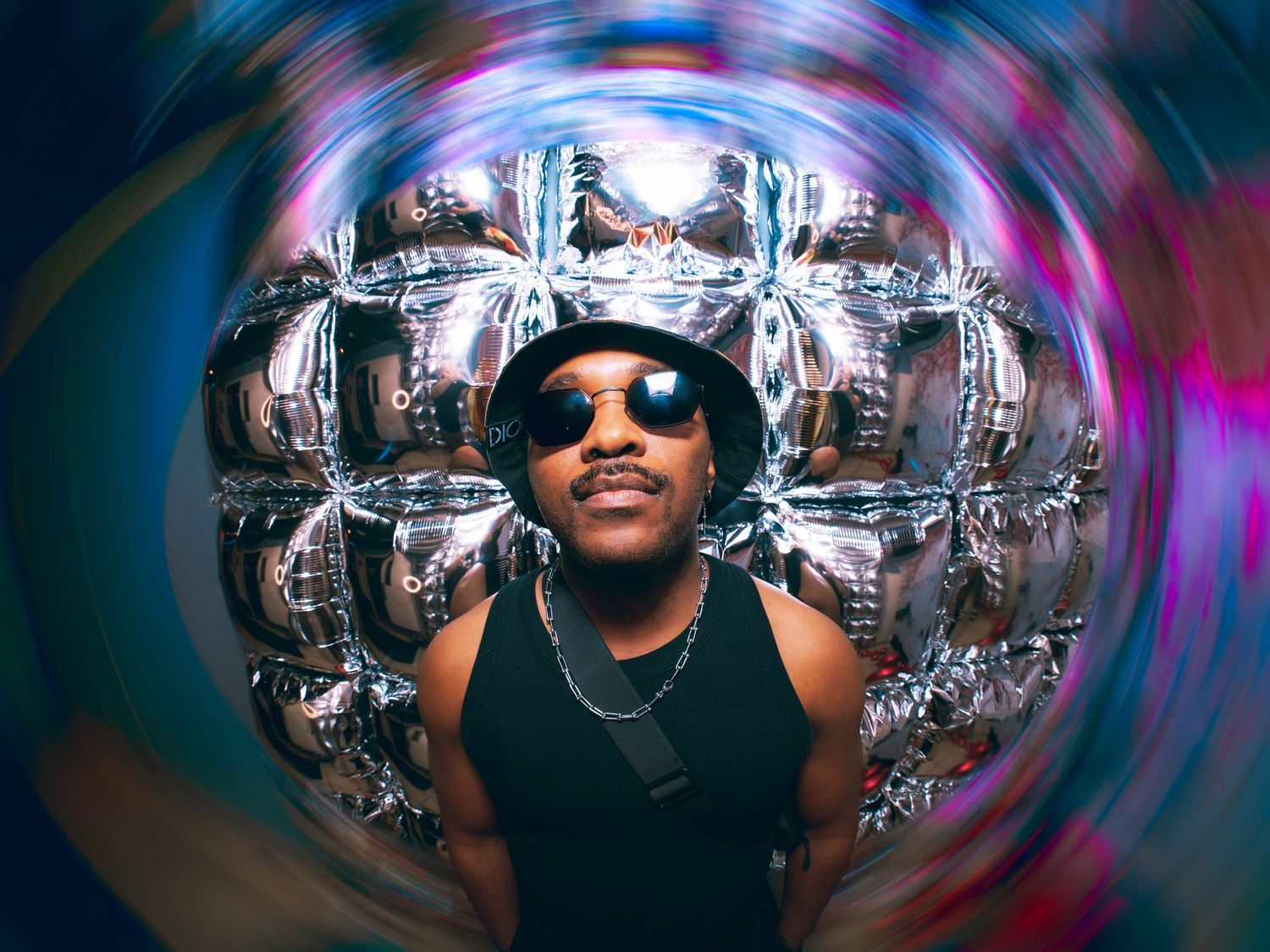 Man wearing bucket hat and sunglasses with swirling silver balloon art in colorful psychedelic background.
