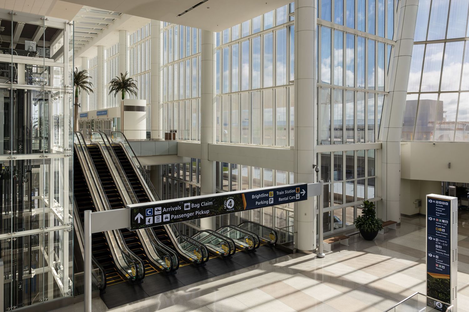 Modern airport terminal interior with escalators, palm trees, and large windows in a bright, spacious atrium.