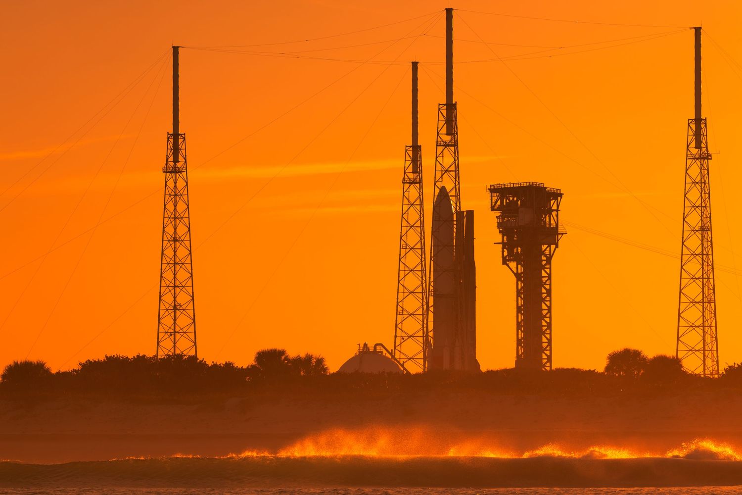 Rocket on launch pad silhouetted against vibrant orange sunset sky at Cape Canaveral space center.