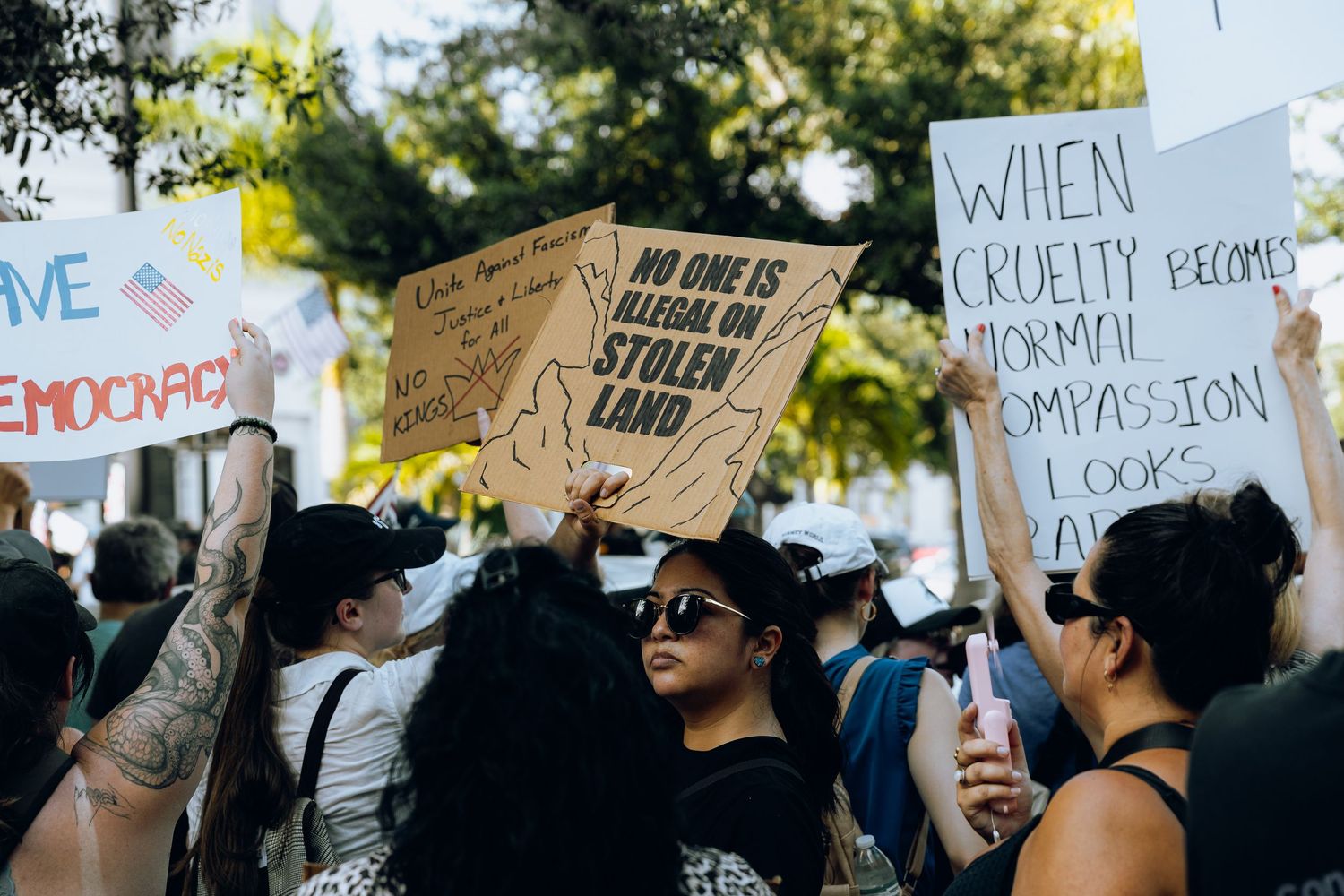 Protesters holding signs at a demonstration with one sign visible against a backdrop of trees and sunlight.