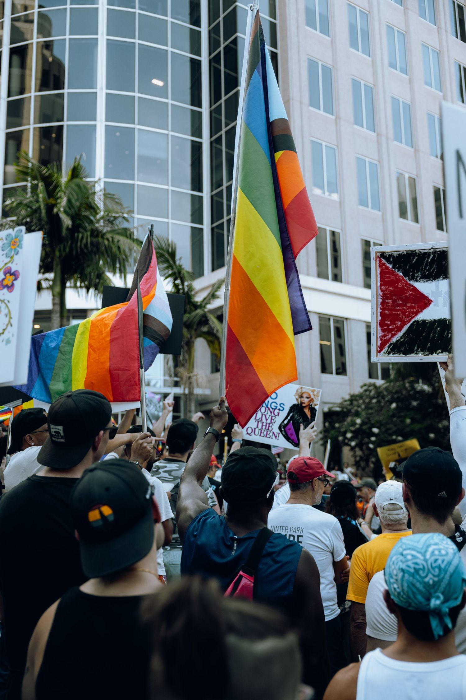 Crowd gathering at protest with rainbow pride flags against modern building backdrop.