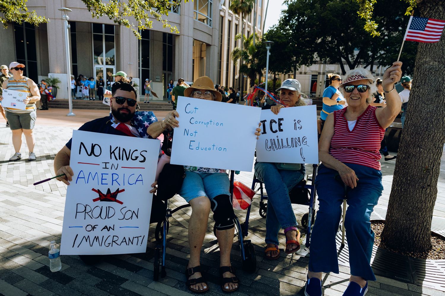 Protesters hold signs at a street demonstration with various political messages while an American flag waves in the background.