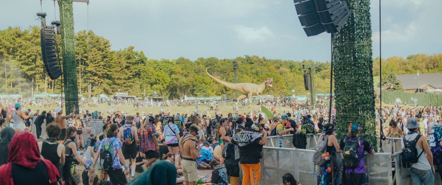 Large crowd of festival attendees gathered in front of outdoor concert stage with sound equipment and green lighting rigs.