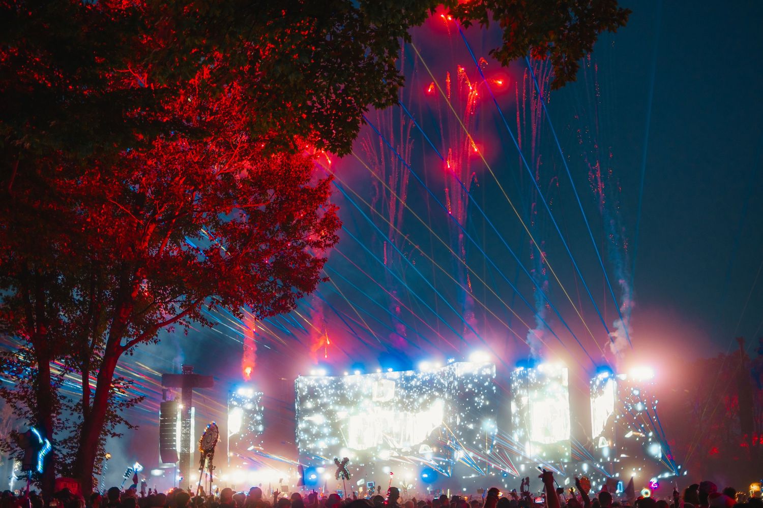 Outdoor music festival stage illuminated with vibrant red and blue lights shining through trees at night.