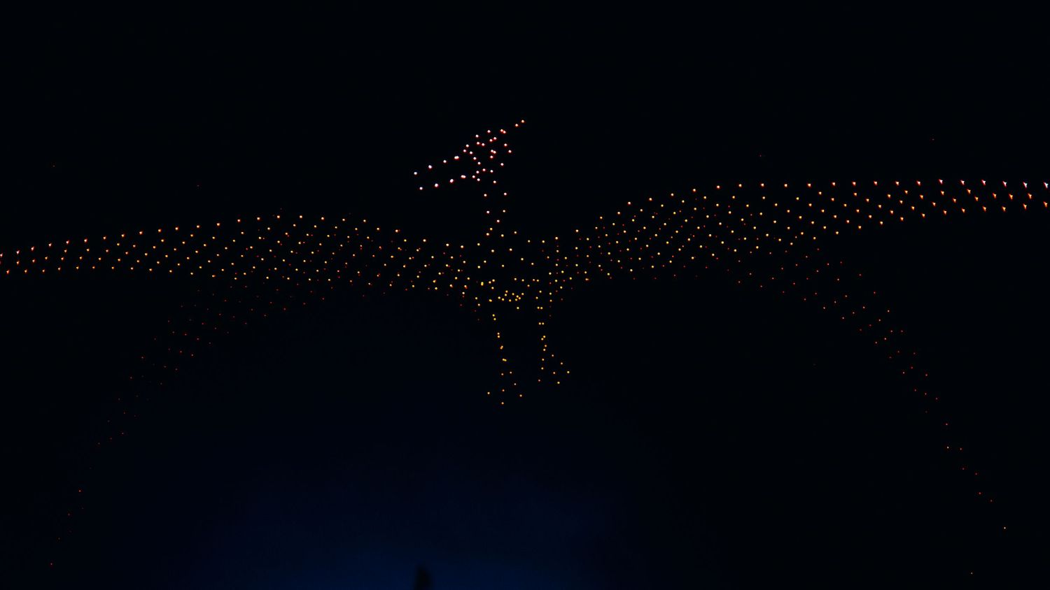 Silhouetted crowd with raised hands at a concert venue illuminated by red and orange stage lights.