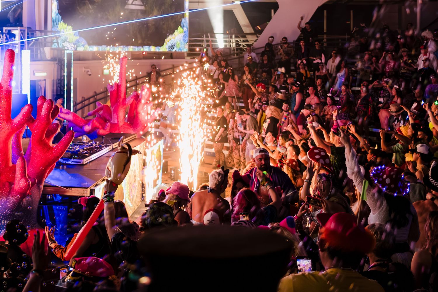 Concert crowd cheers as pyrotechnic effects and colorful lights illuminate a packed indoor music venue during performance.