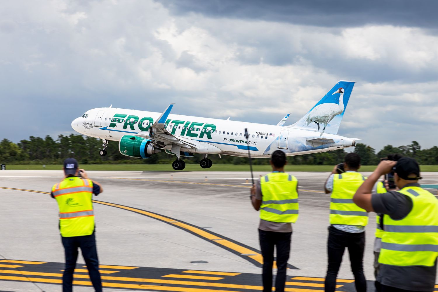 Frontier Airlines Airbus A319 taking off on runway with ground crew in yellow vests photographing the departure.