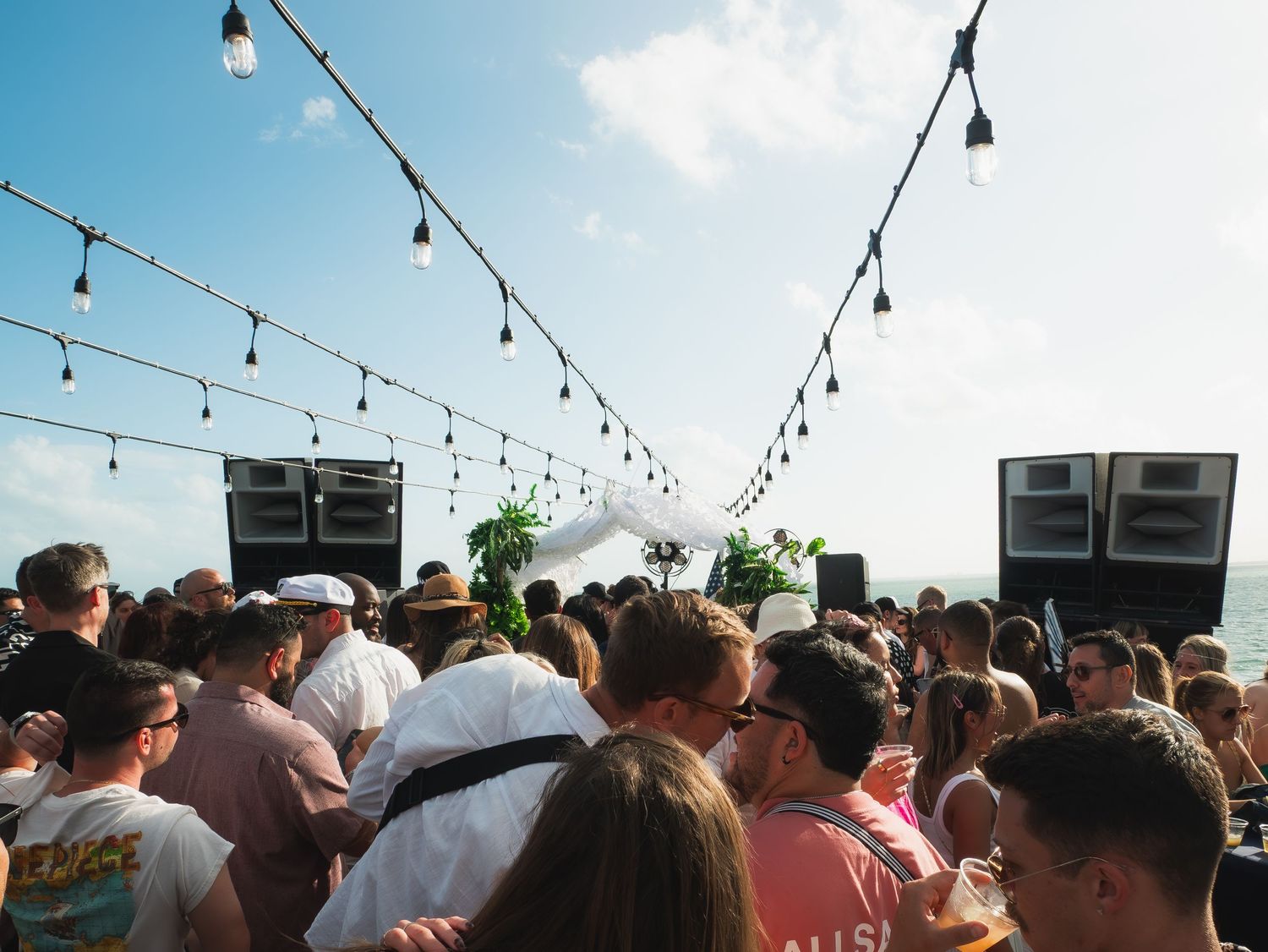 Crowd gathered at an outdoor rooftop party under string lights with large speakers on a sunny day.