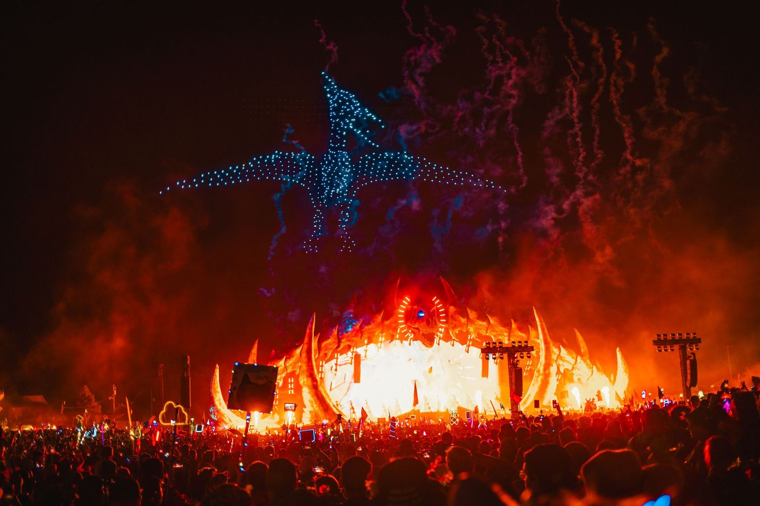Glowing blue dinosaur sculptures illuminate a dramatic nighttime display with fire and smoke against a dark landscape.