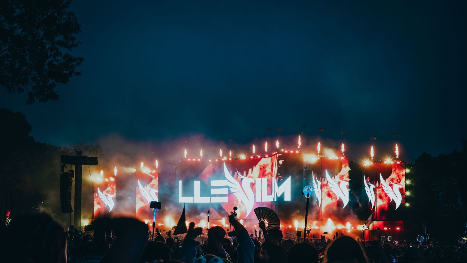 Concert crowd silhouettes against a vibrant red stage lighting backdrop during a nighttime outdoor performance.