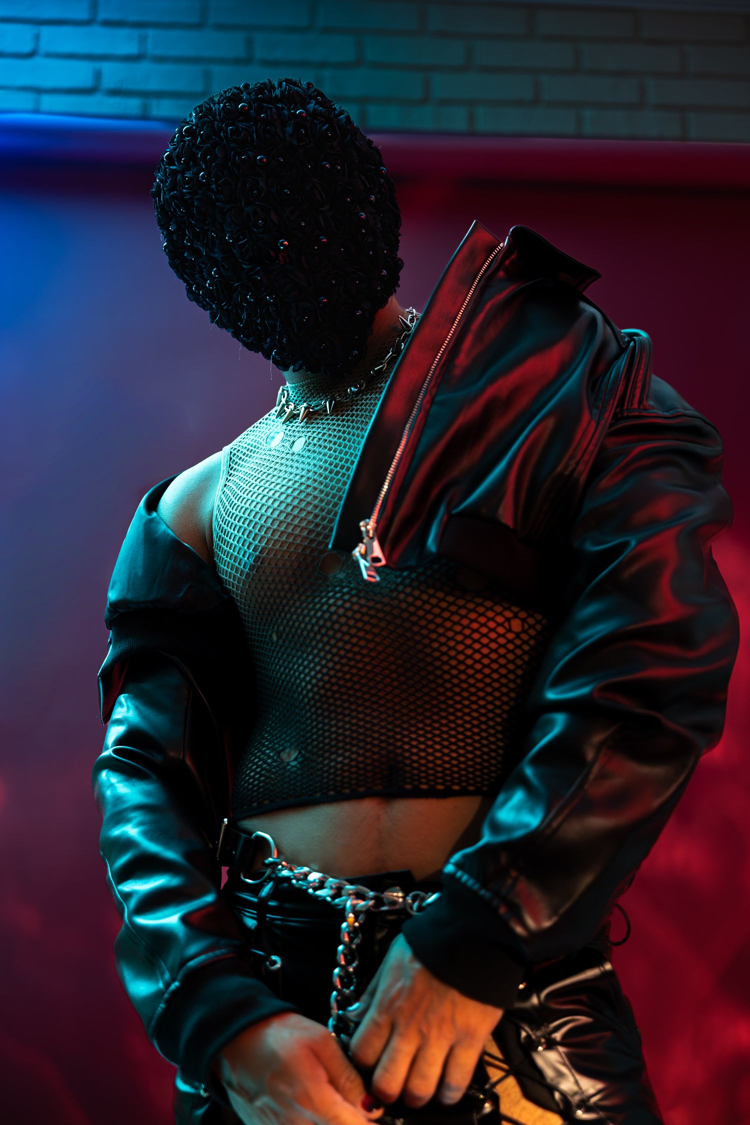 Person in black leather jacket, mesh top, chain belt, and beaded hat poses under dramatic red and blue neon lights.