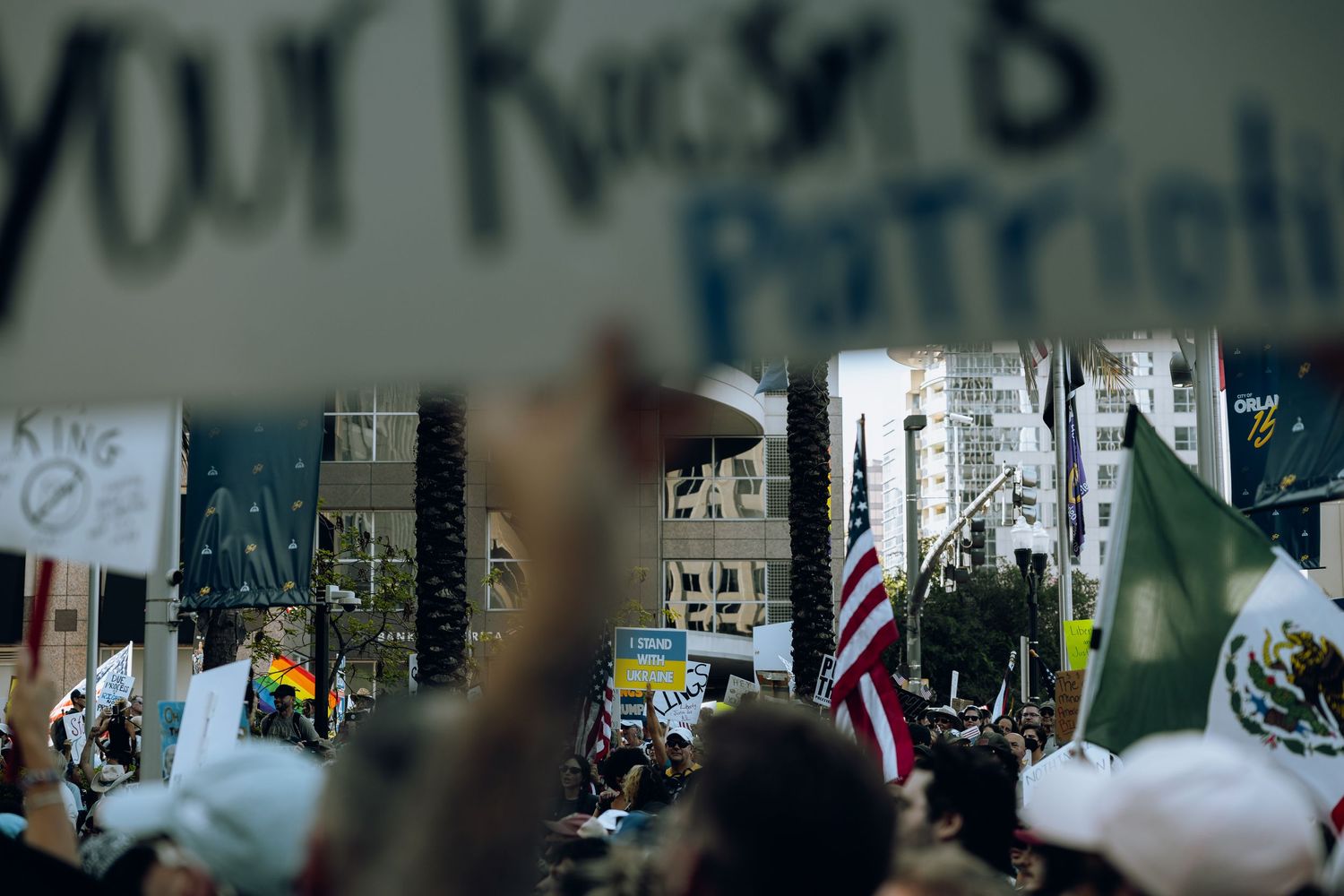 A large crowd of protesters gather at a rally with raised signs and flags visible against urban buildings.