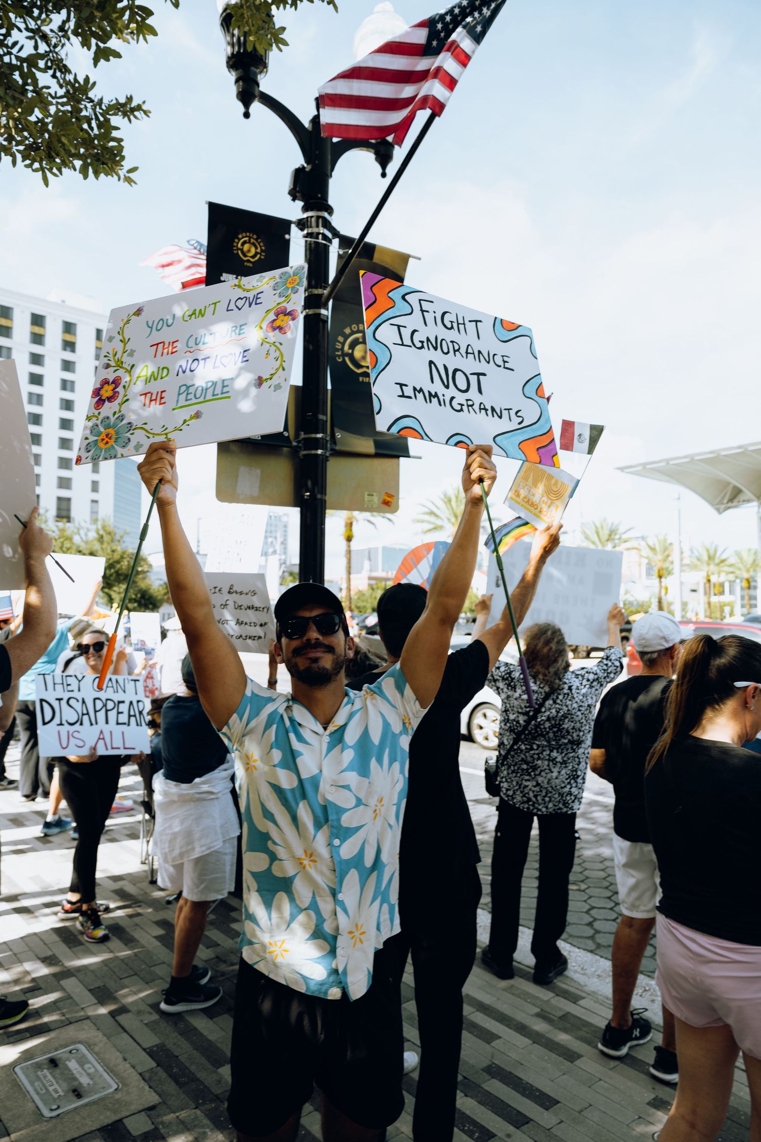 Crowd of demonstrators waving American flags and protest signs at a rally near urban buildings and palm trees.