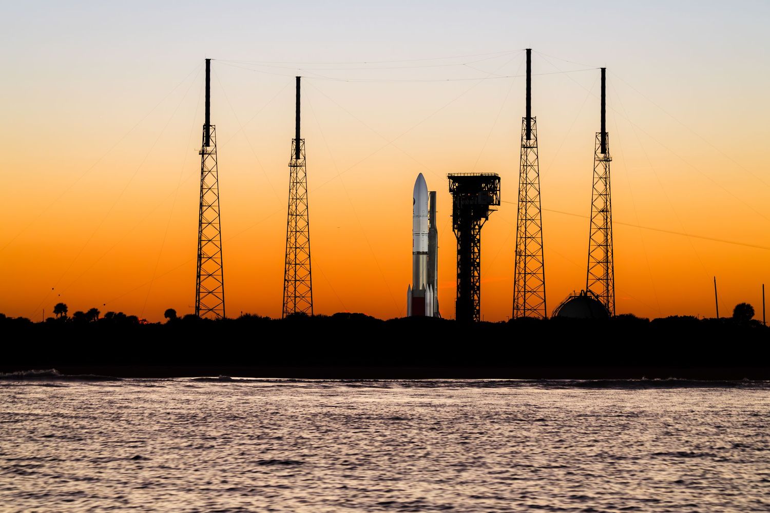 A rocket on a launch pad silhouetted against a vibrant orange sunset sky, reflected in calm water below.
