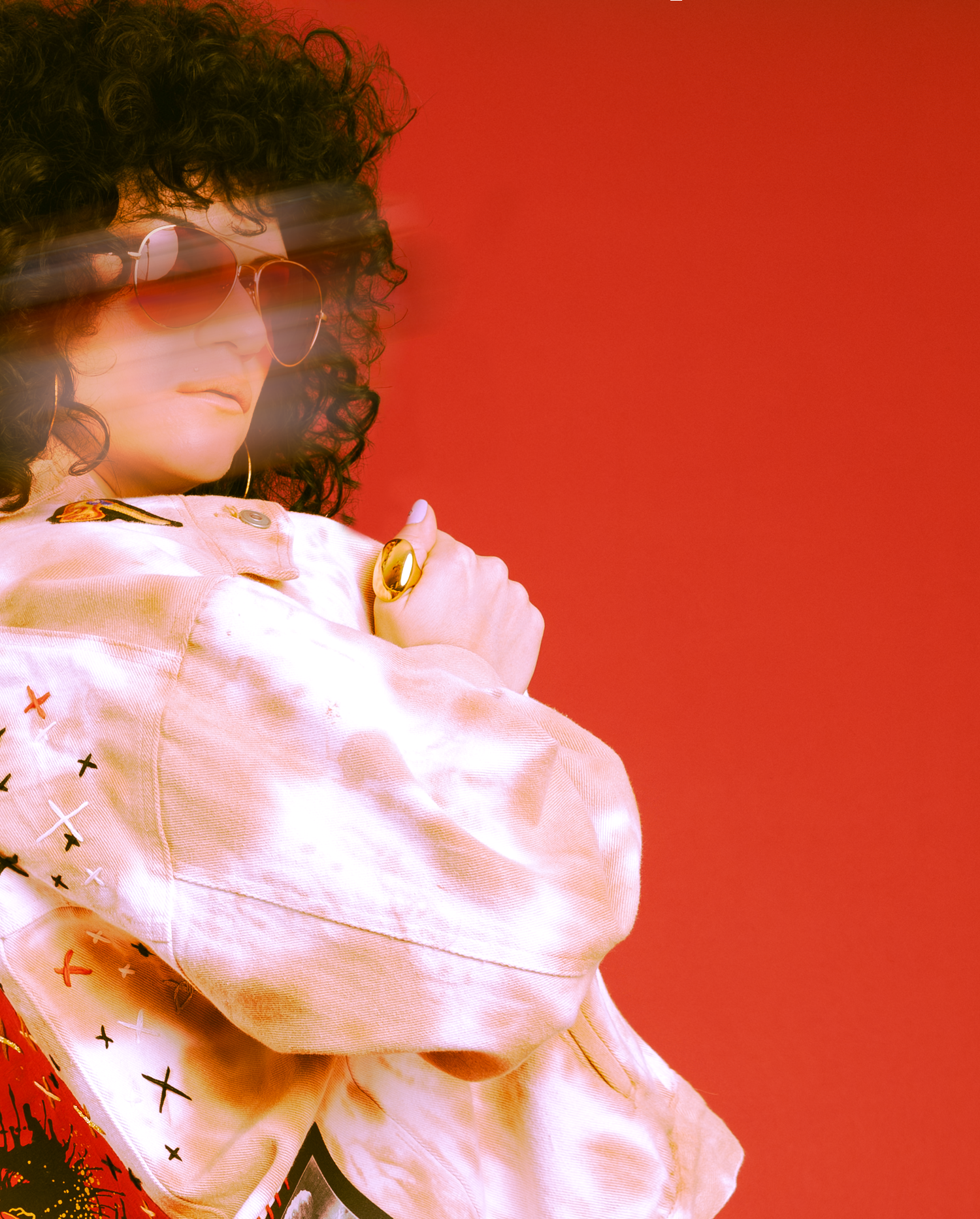Person with curly hair and sunglasses wearing a white jacket with X embroidery against a red background.