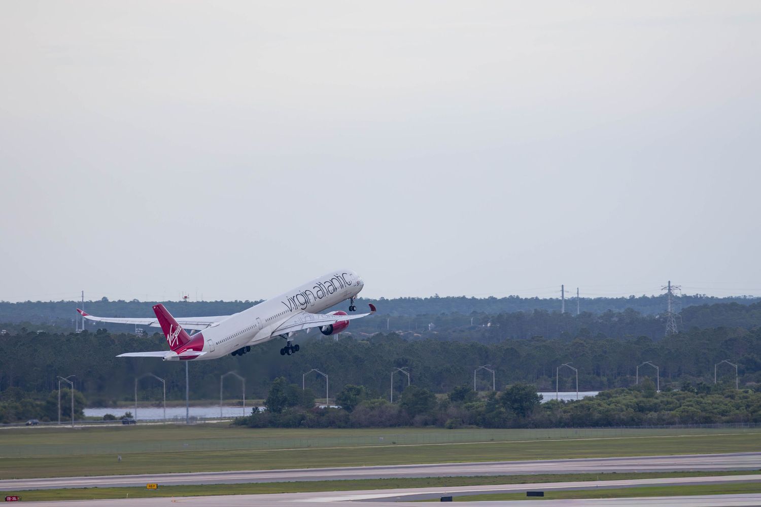 Virgin Atlantic airplane taking off from a runway surrounded by lush green trees under a cloudy sky.