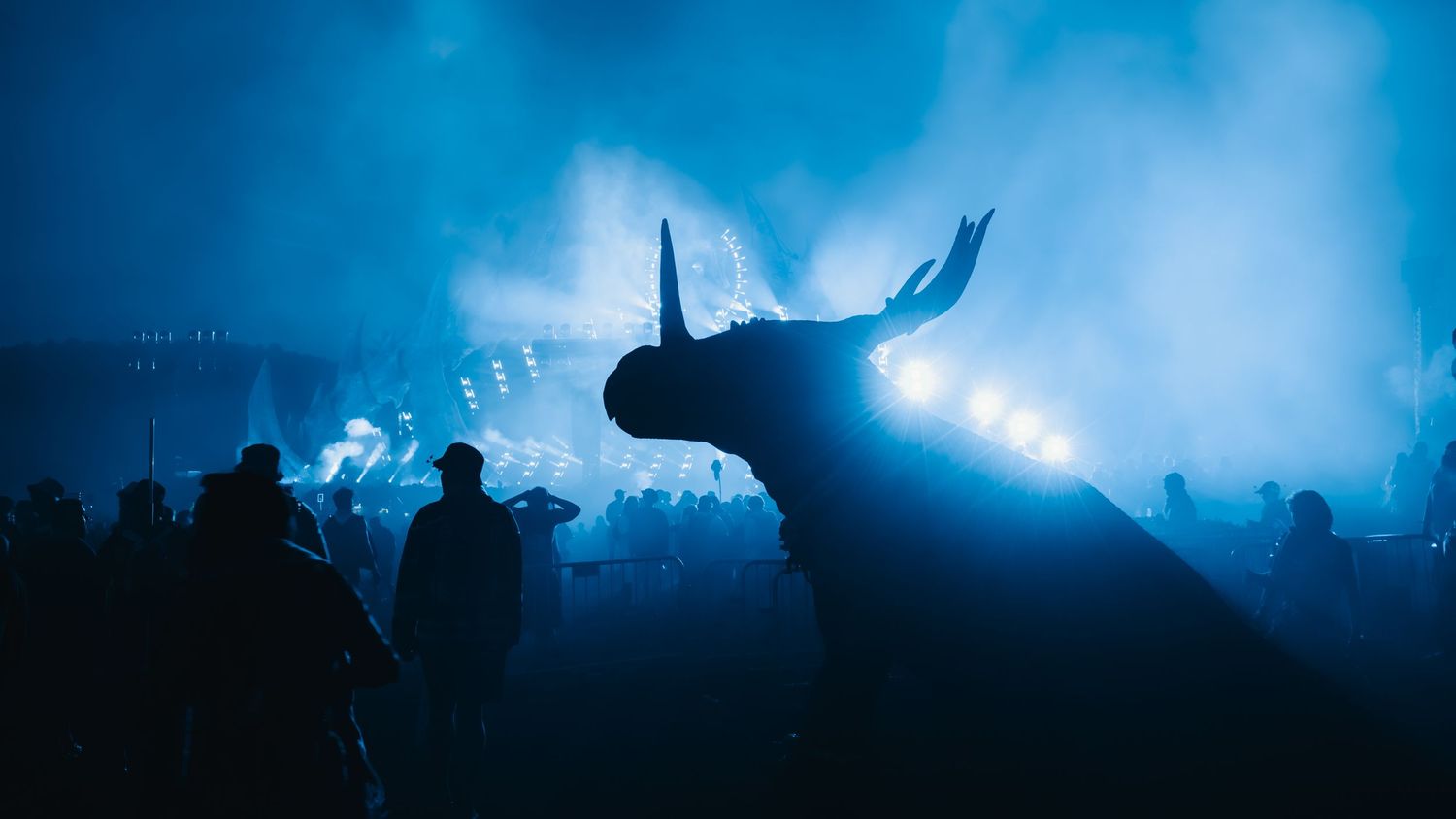 A dramatic blue-lit silhouette of a large bull sculpture looms over a crowd at a nighttime festival event.