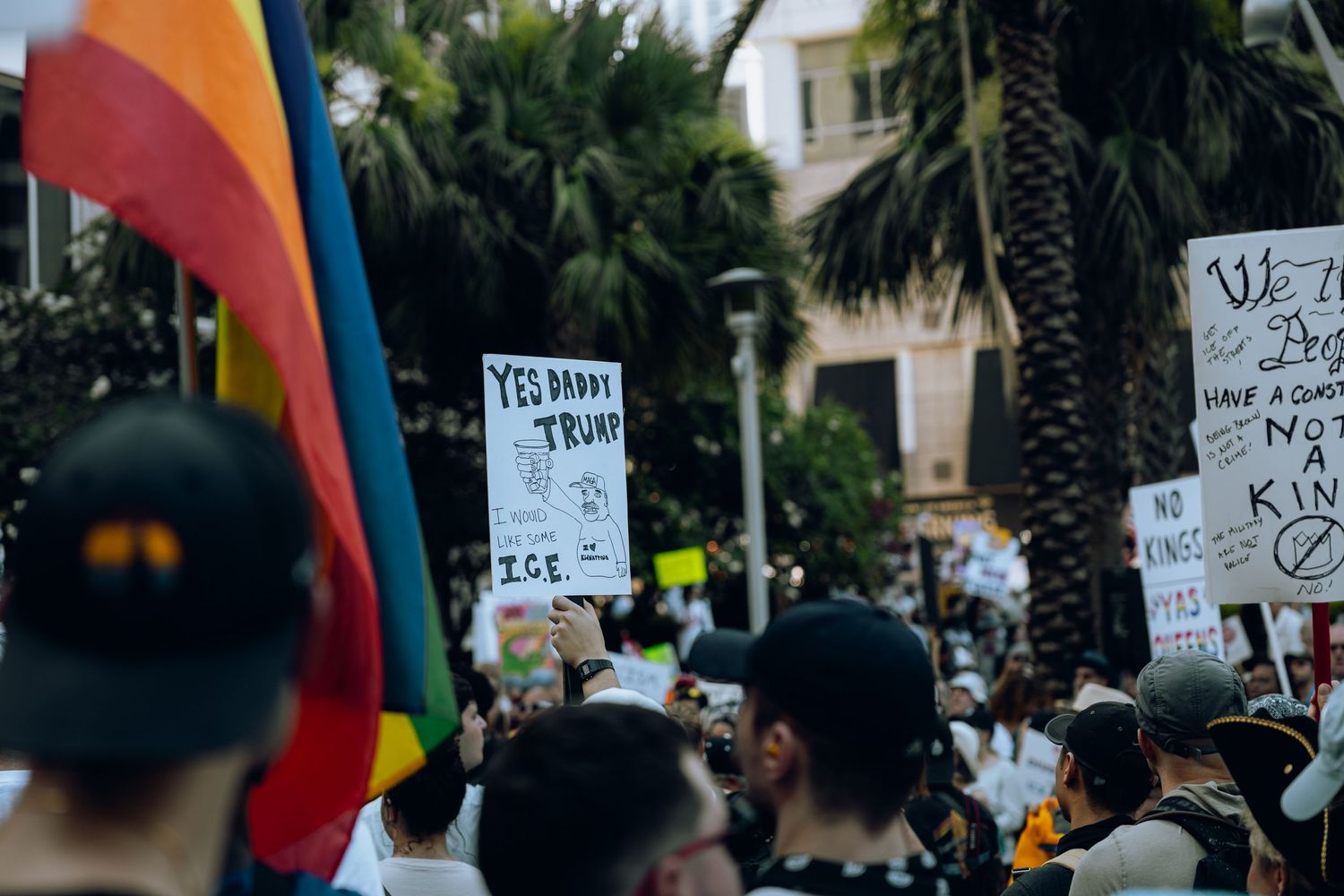 Protesters hold signs while gathering at a rally with rainbow pride flag visible in the foreground near palm trees.