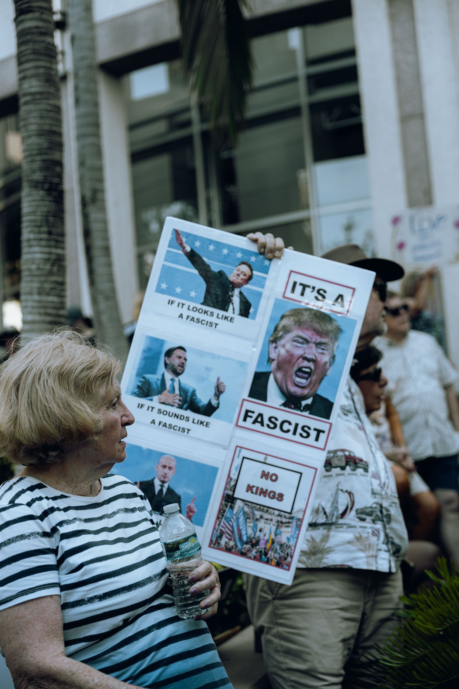 Protester holds sign with anti-Trump messages at outdoor political rally near palm trees.