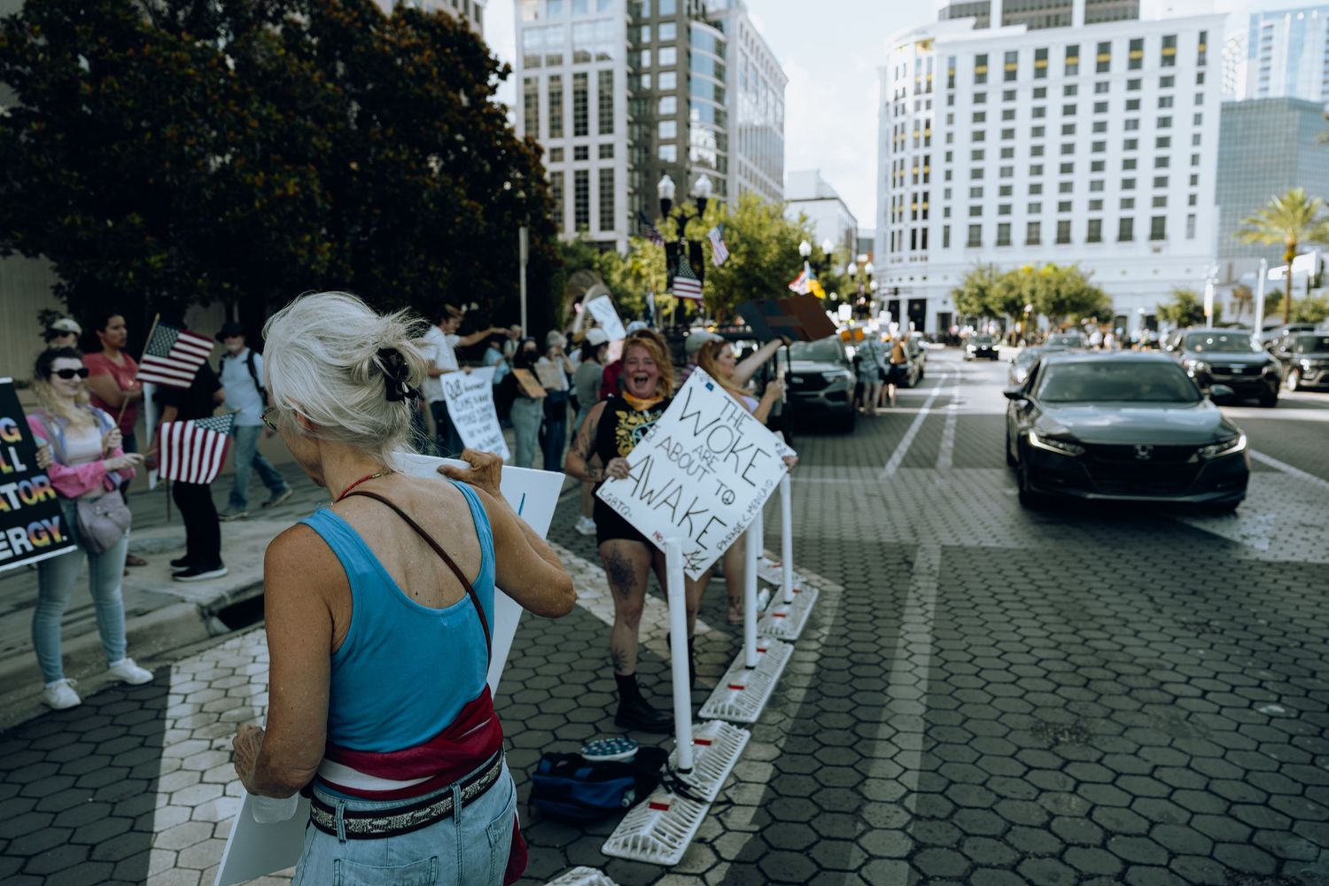 Protesters line a city street with signs while tall buildings and trees fill the urban backdrop.
