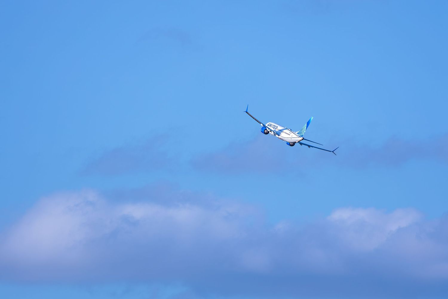 A commercial airplane ascending through a clear blue sky with soft white clouds below.