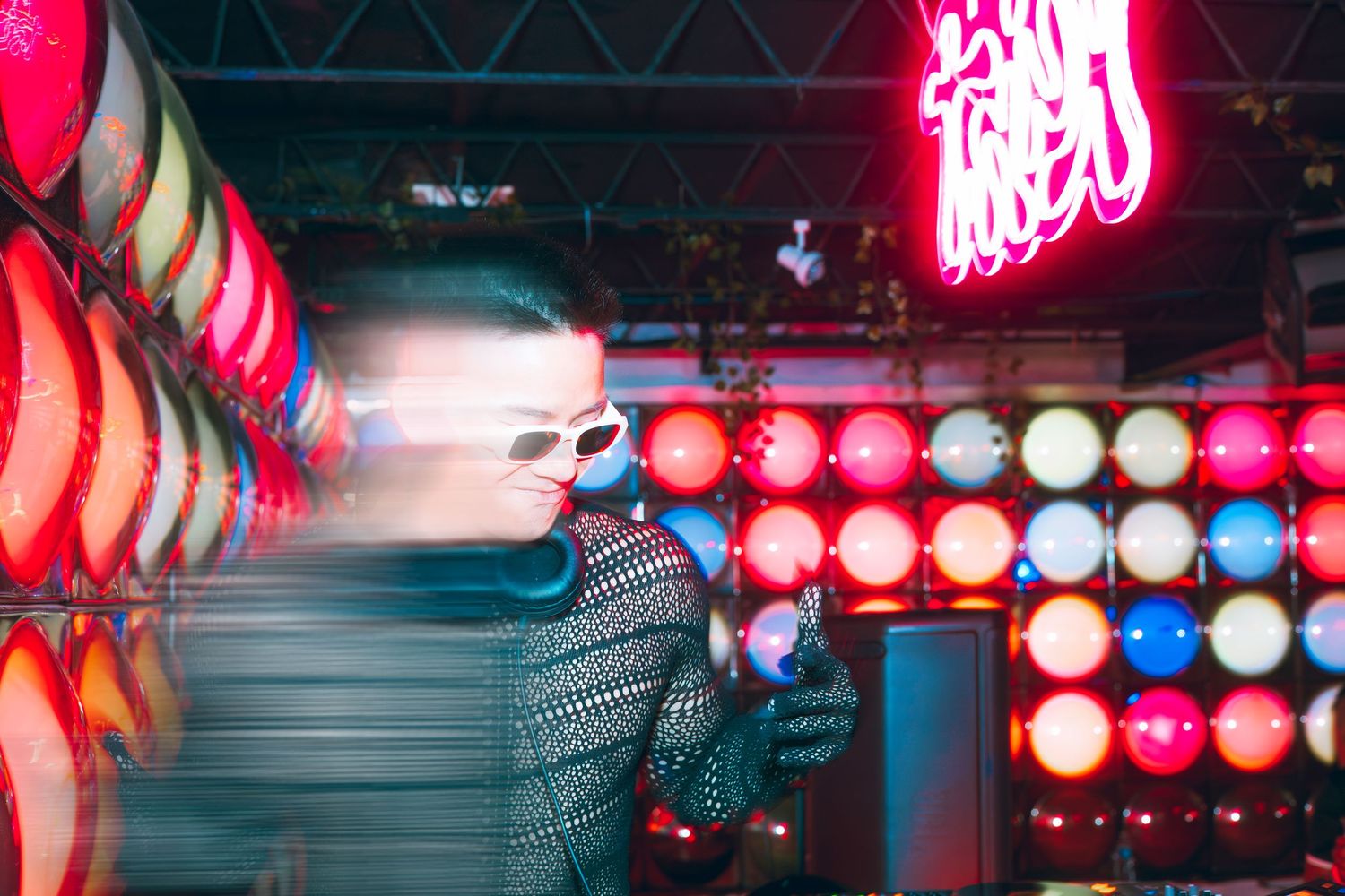 Person wearing white sunglasses posing beside glowing neon lights and illuminated orbs at night.