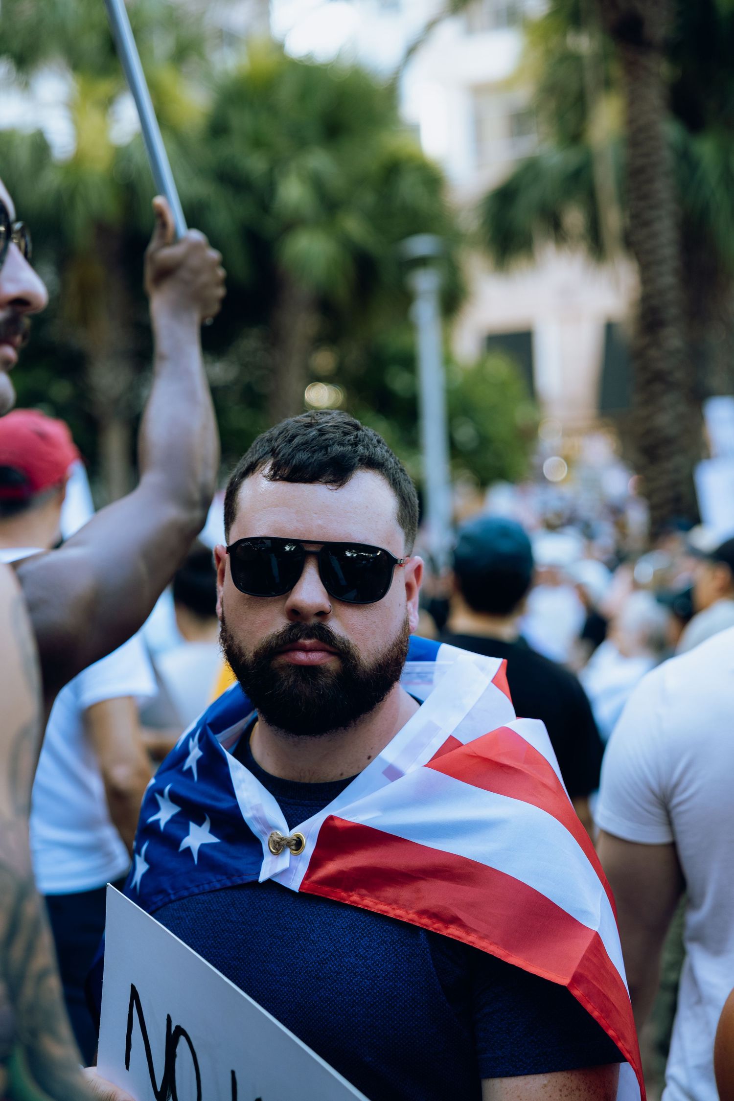 Bearded protester wearing sunglasses and American flag at political demonstration.