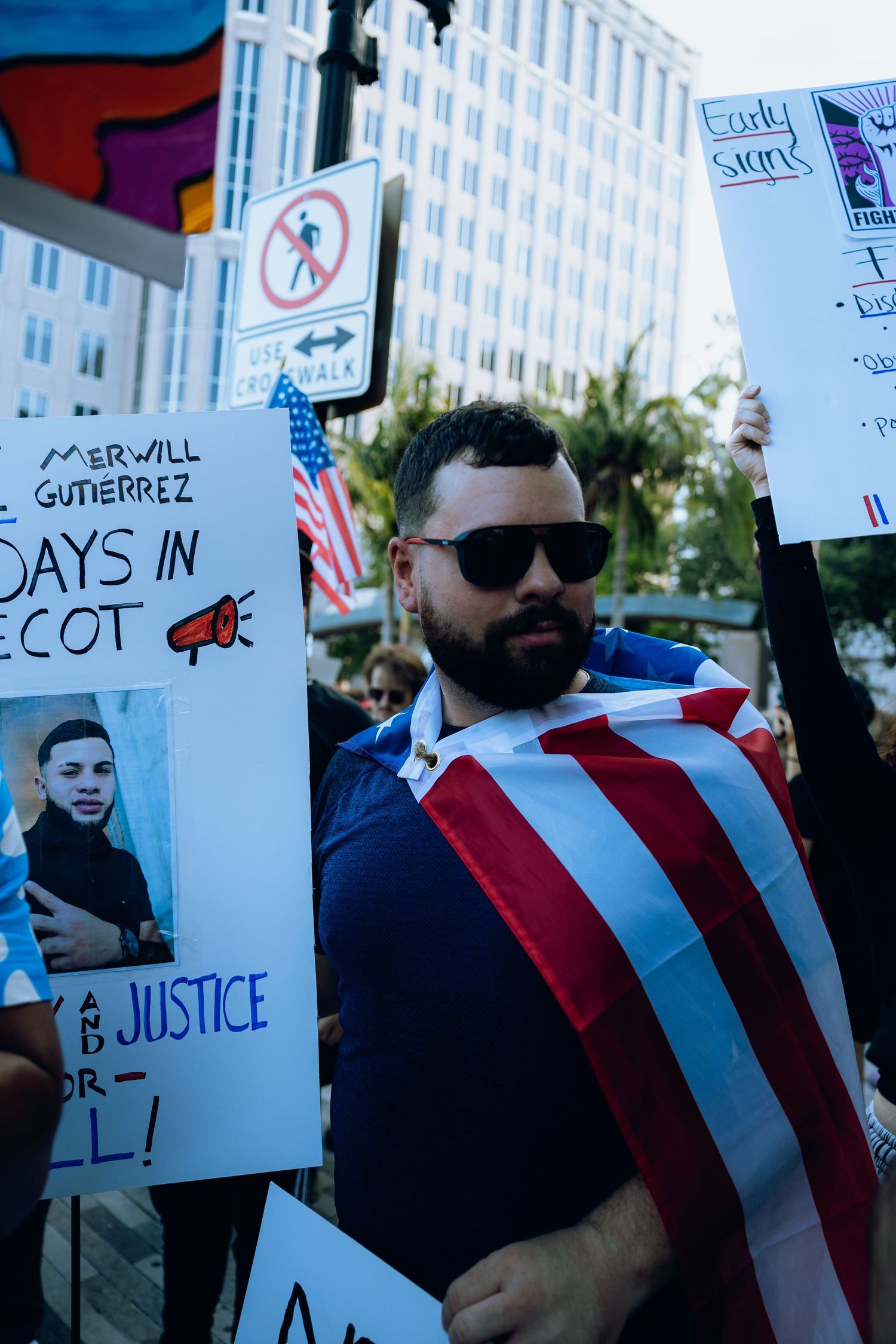 Protestor draped in American flag standing next to signs at urban demonstration.