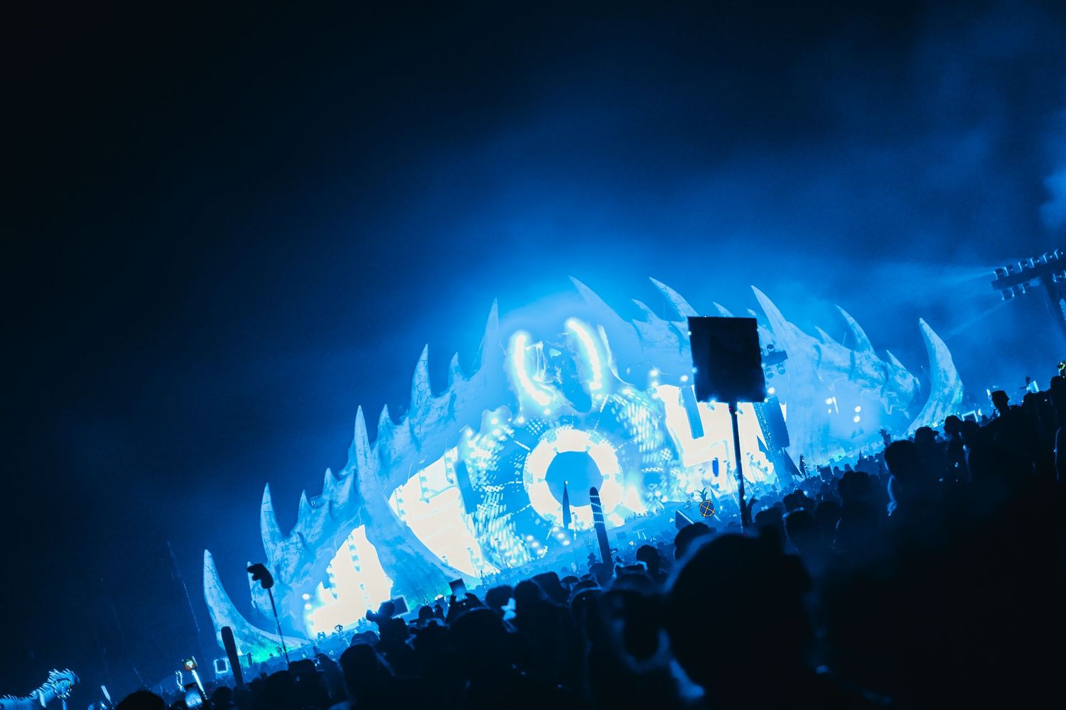 A panoramic view of a concert crowd silhouetted against bright blue stage lighting and visual effects.