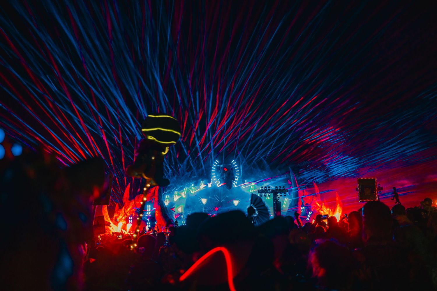 A colorful light show illuminates a dark outdoor concert venue with silhouettes of people and laser beams in the sky.