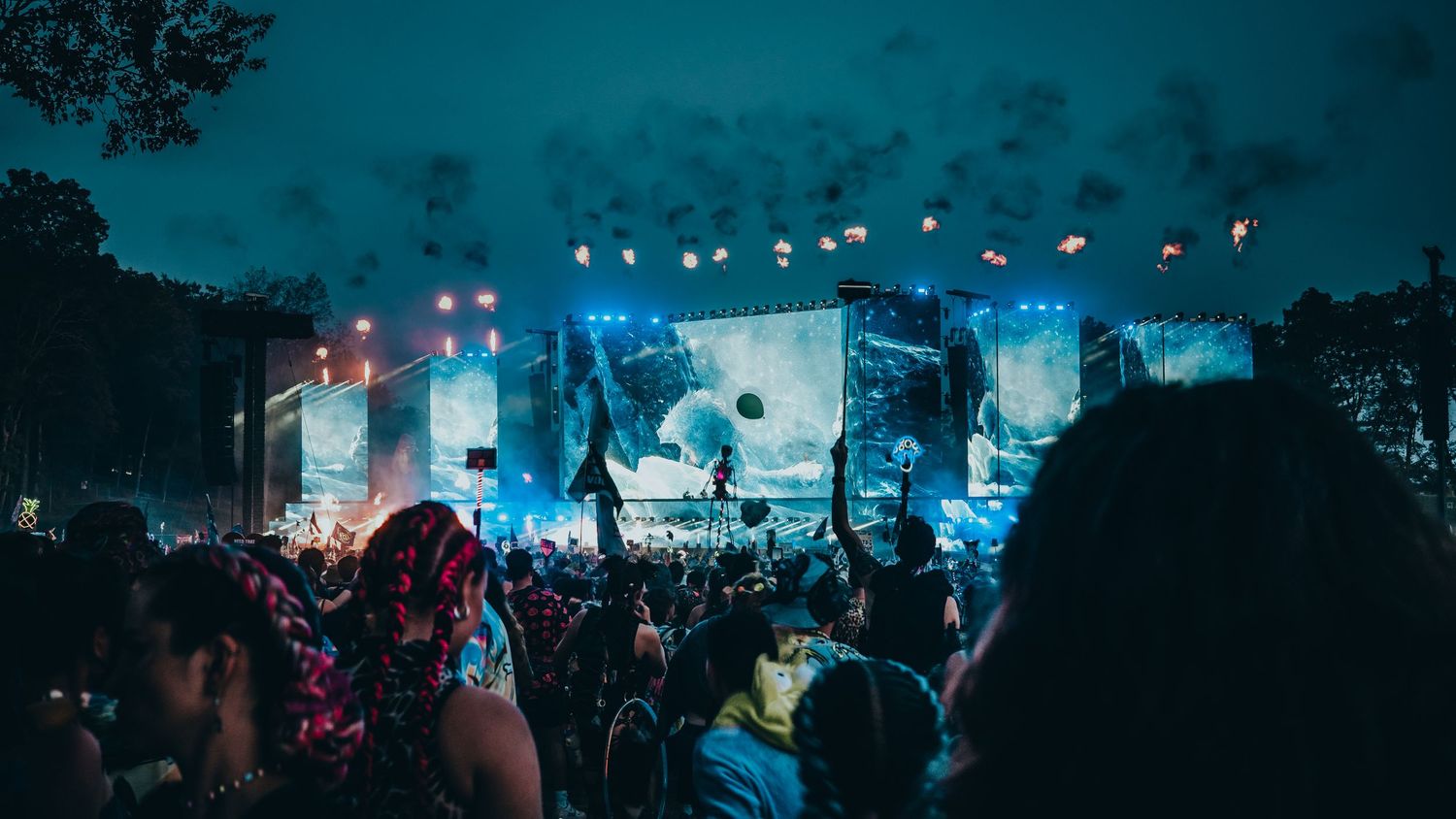 Silhouettes of concert crowd enjoying outdoor music festival with bright stage lights illuminating the night sky.