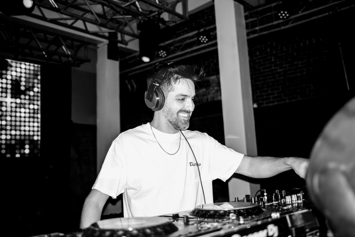 DJ performs on stage behind mixing equipment at a nightclub with dark lighting and atmosphere.