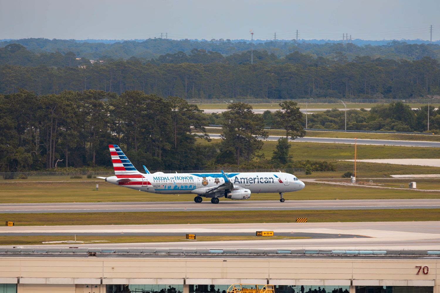 American Airlines Airbus A321 aircraft with special livery taxiing on runway at a sunny airport with green trees in background.