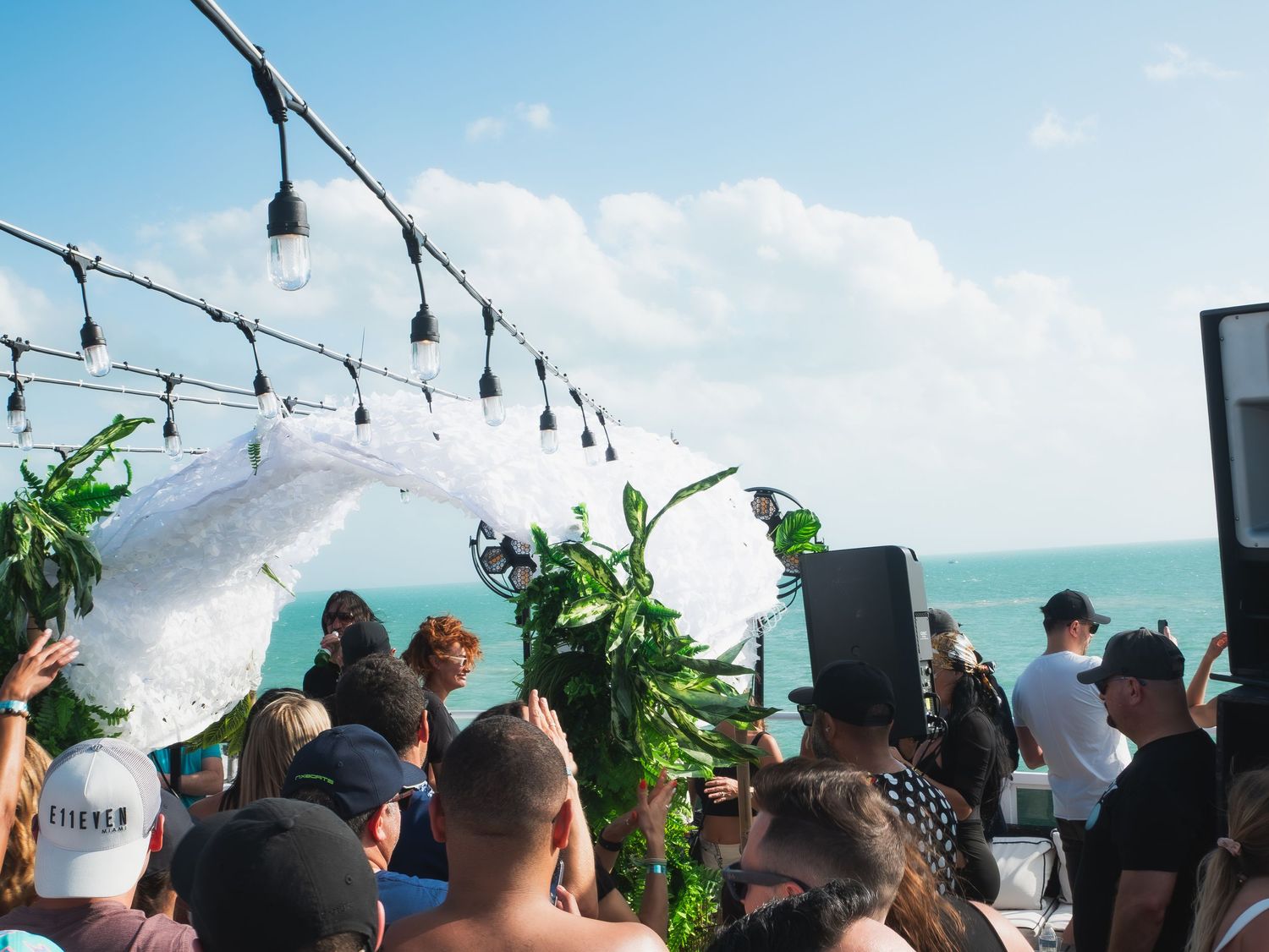 Beachside event setup with string lights and tropical decor overlooking turquoise ocean waters during daytime gathering.