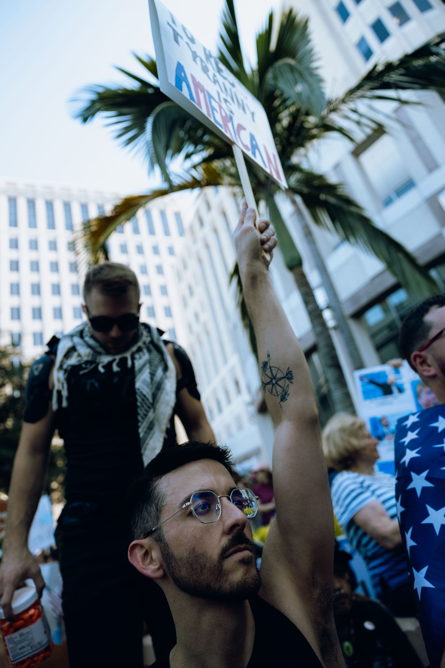 A protester in a crowded city raises a sign during a demonstration with palm trees and buildings in the background.