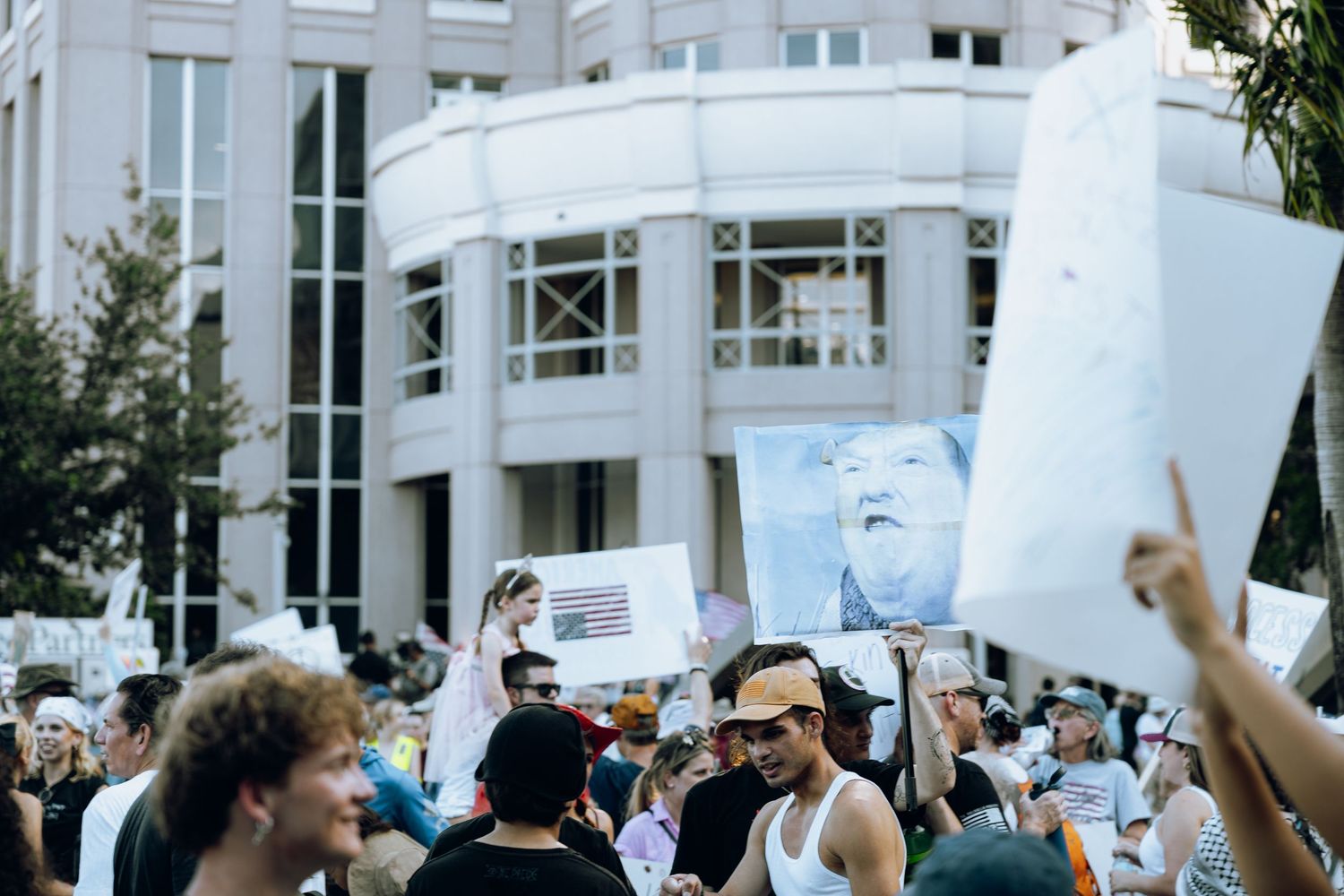 Protesters holding signs and banners at a demonstration outside a modern white building.
