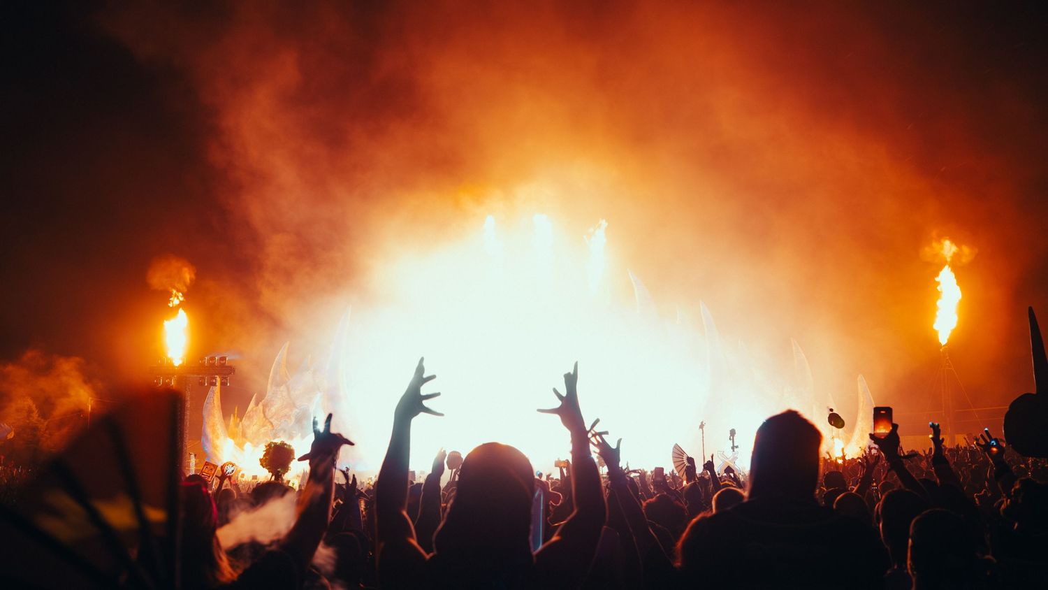 Silhouetted crowd with raised hands at a concert with dramatic orange lighting and smoke effects in the background.