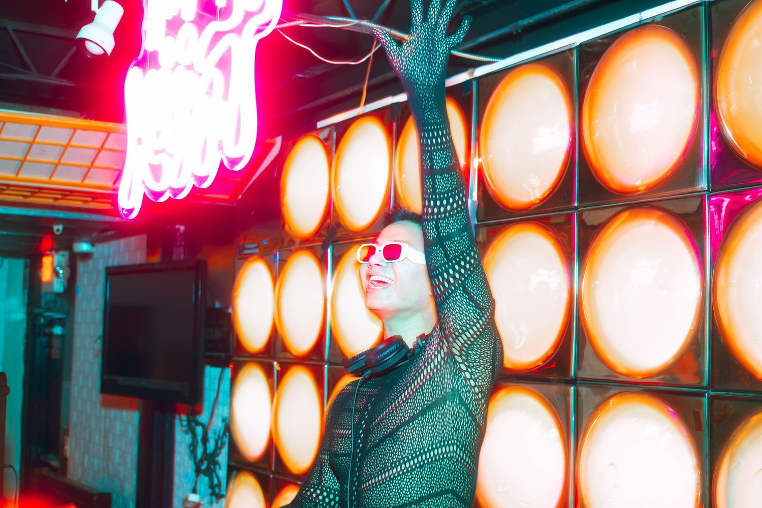 A performer in a mesh outfit and pink sunglasses poses dramatically in front of large round orange lights at a nightclub.
