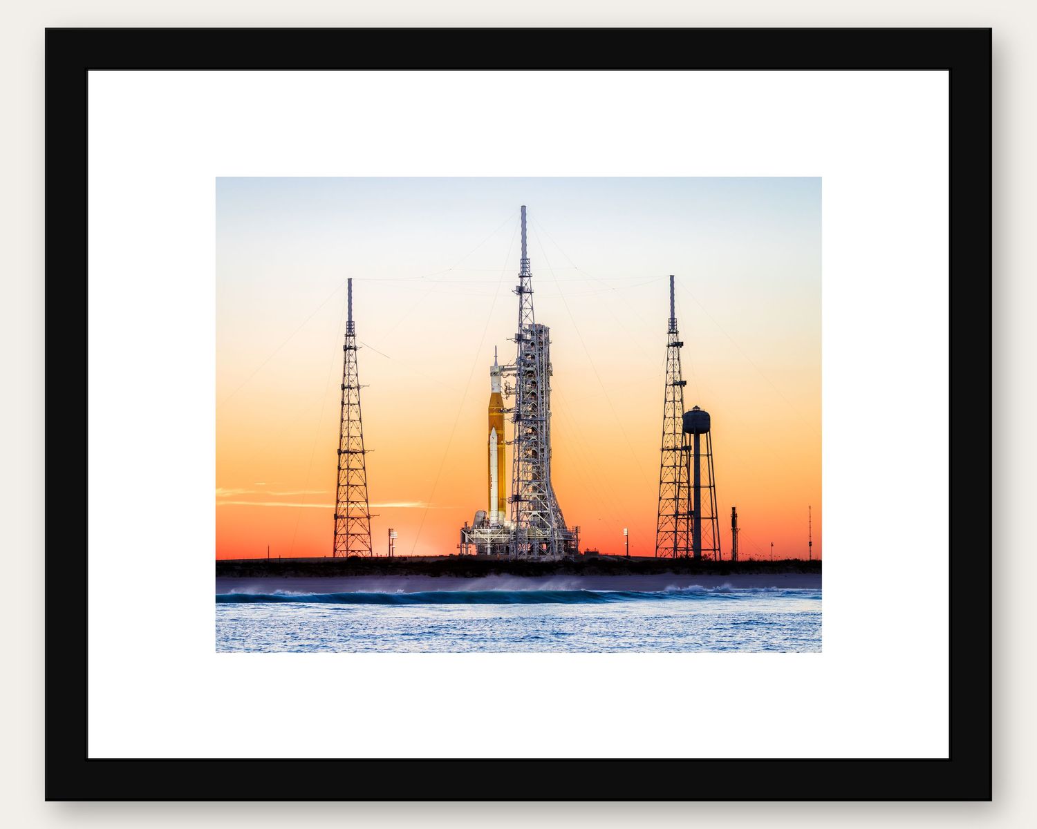 Framed print of a NASA rocket on launch pad at sunset with orange sky, displayed with white mat border.