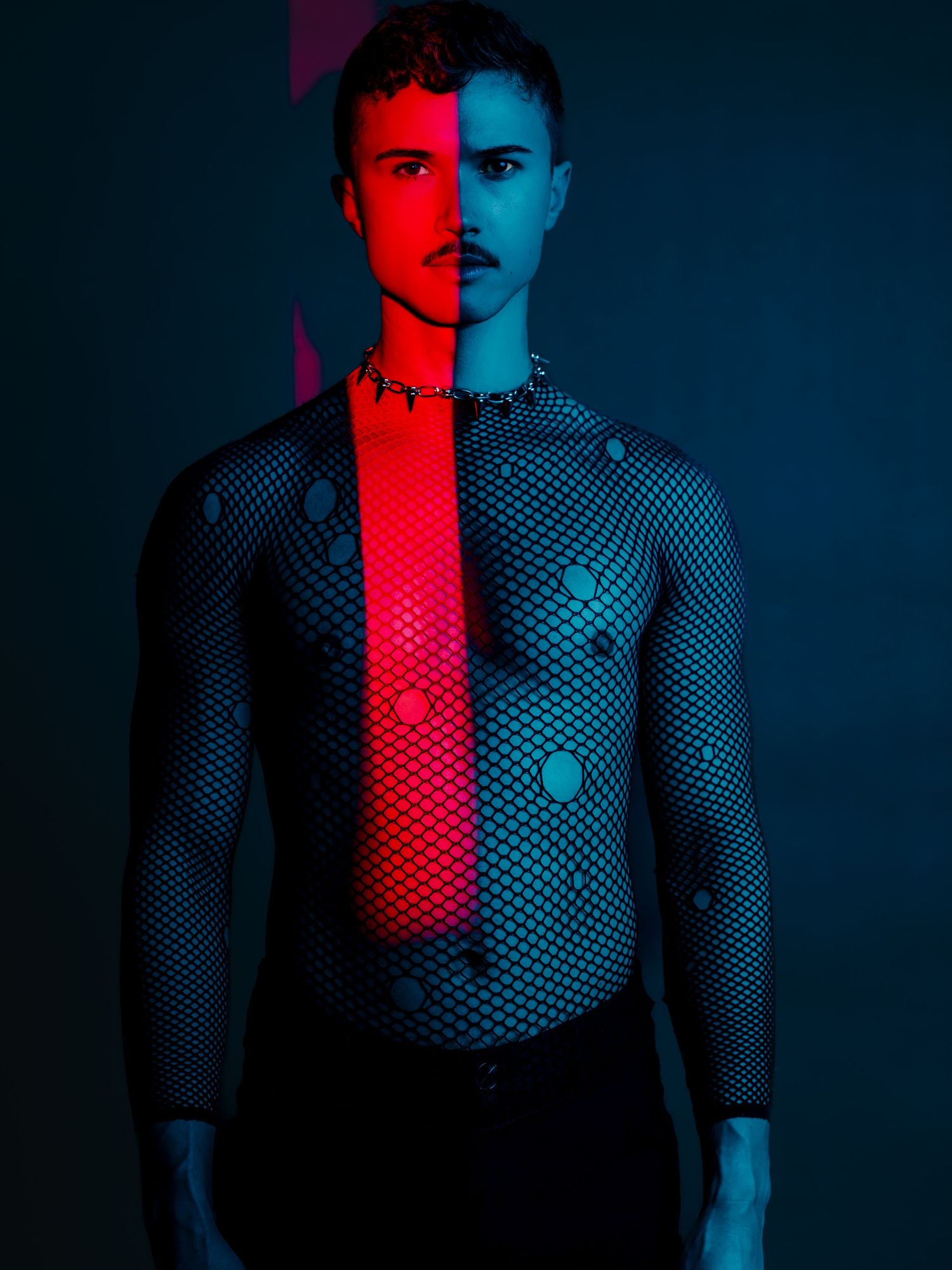 Man in mesh long sleeve top illuminated by red and blue neon lights in dark studio.