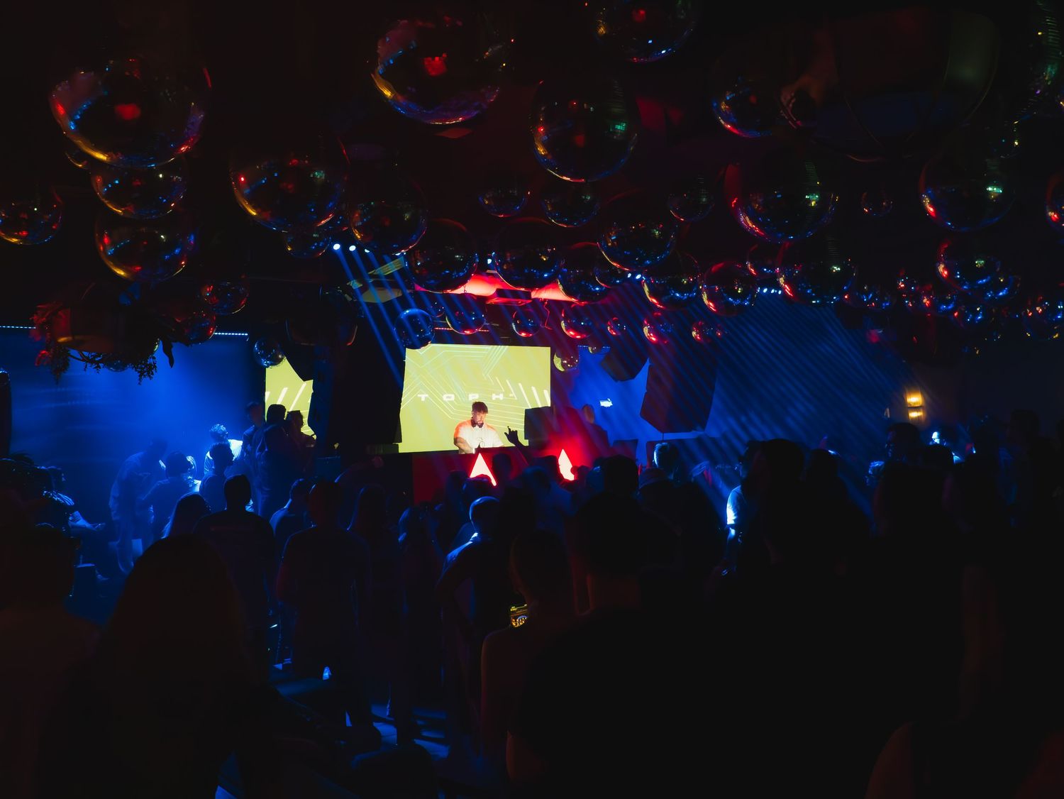 Dark nightclub with silhouetted crowd, blue lighting, and projection screens visible in a dimly lit atmosphere.