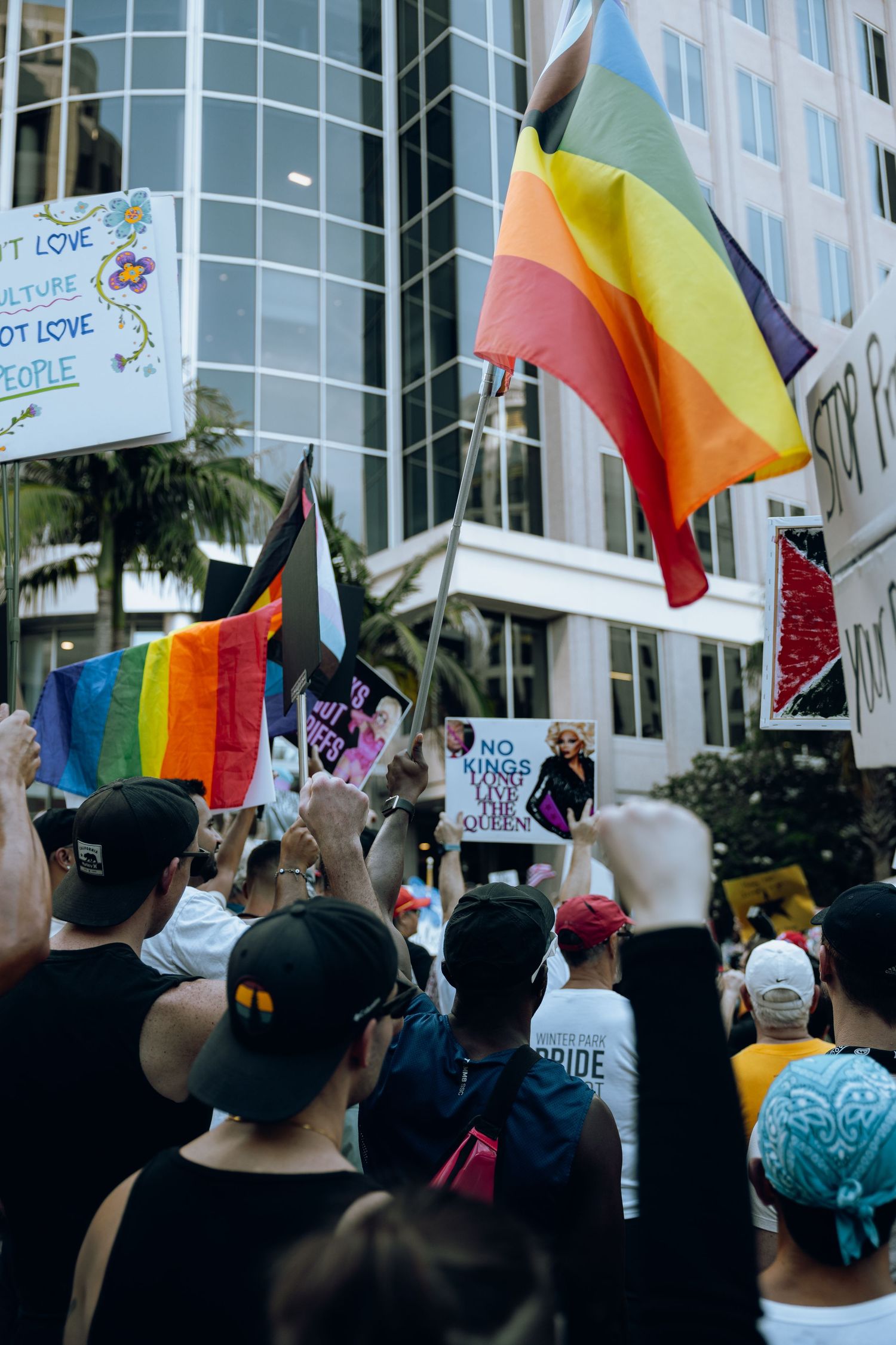 Pride flags wave above crowd during protest outside modern office building.