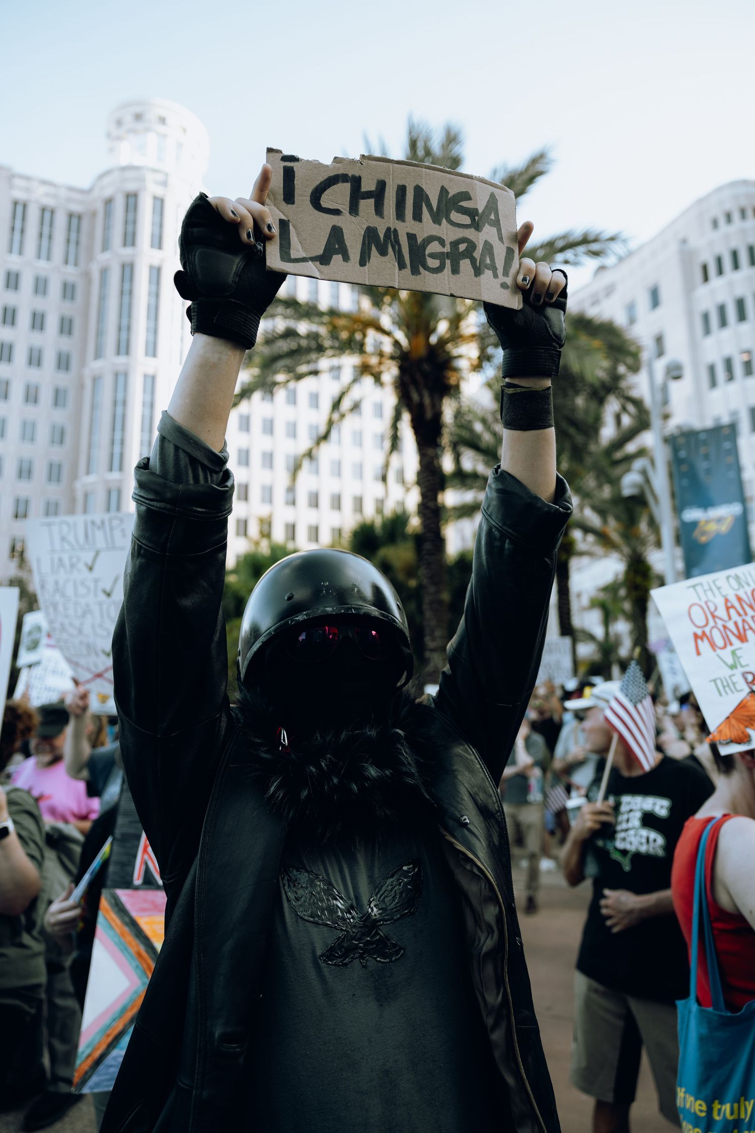 Protestors holding signs at a demonstration with palm trees and buildings visible in the background.